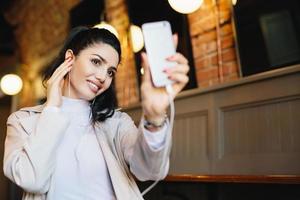 mujer atractiva con cabello oscuro, piel sana y ojos brillantes sosteniendo un teléfono inteligente en la mano haciendo selfie y escuchando música o audiolibros mientras descansa en un café. concepto de personas y ocio foto