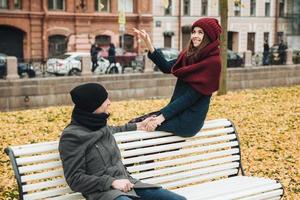 Beautiful female and male have fun outdoors, sit on bench, keeps hands together, look at each other with great love, being very romantic and relaxed. Togetherness concept photo