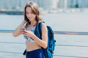 Horizontal portrait of lovely female with perfect figure, wears leggings and top, has focused look into smart phone, carries big rucksack, tries to find correct route while covers long distance alone photo