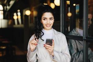 Horizontal portrait of brunette adorable woman in white clothes using her cell phone while standing outside having good mood touching her black curl of pony tail trying to make decision. Elegant girl photo