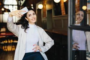 Waist up portrait of beautiful lady with dark hair tied in pony tail wearing white jacket sitting in cafe making selfie using her modern smartphone and listening to music with earphones. People, rest photo