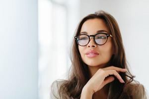 Close up shot of delicate woman wears optical glasses with dark frames, touches gently chin, looks aside thoughtfully, has dark straight hair, copy space on left side. Thoughtful businesswoman photo