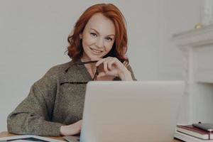 Successful professional businesswoman with red hair works remotely on laptop computer, reads article about business affairs online, sits at desktop, uses modern technologies, prepares financial report photo