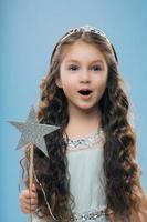 Small little child female princess wears crown and dress, holds magic wand, has long dark curly hair poses over blue background. Beautiful kid preapres for carnival or festive event. Childhood concept photo