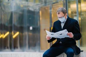 una foto al aire libre de un jefe serio se toma un descanso después de caminar, lee el periódico, usa anteojos para una buena visión, máscara médica para protegerse de los virus, descubre noticias sobre la propagación del coronavirus