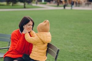 Positive young brunette mother wears red sweater, has fun together with her little nephew who touches her cheeks, dressed in jacket and hat, pose outside against blurred green grass, sit on chair photo