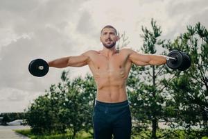 Strong handsome muscular bodybuilder pumps up arm muscles with barbells, does bidybuilding biceps exercises outdoor, poses with naked torso against green trees and sky, makes weight exercise photo