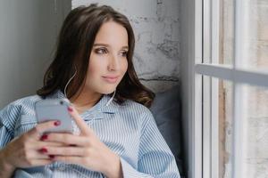 una mujer morena pensativa escucha un audiolibro, sostiene un teléfono inteligente moderno en las manos, disfruta de un sonido perfecto en los auriculares mira pensativamente a la ventana vestida con ropa informal. la mujer escucha música en la lista de reproducción foto