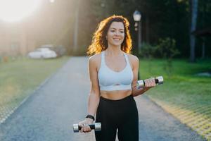 Photo of slim brunette young woman raises dumbbells has morning workout poses against sunrise dressed in cropped top and leggings works on arms muscles smiles positively leads sporty lifestyle