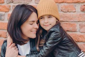 Close up shot of tender pleased young lady smiles gladfully and closes eyes, recieves hug from small daughter who wears leather jackt and hat, pose together over brick wall. Motherhood concept photo