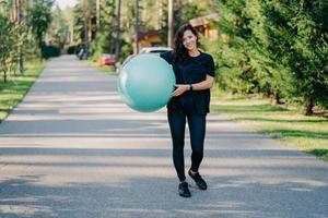 Fitness woman exercises with fitness ball outdoor walks on road near forest has healthy lifestyle dressed in black active wear, has happy expression, stays in good physical shape. Fitness concept photo