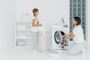 contenta de que la madre y la hija estén ocupadas lavando la ropa en casa, tienen caras felices, la mujer morena carga la lavadora, la niña se para cerca de la canasta con ropa blanca. tareas domésticas, concepto de limpieza. foto