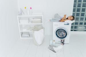 Shot of tired small kid sleeps on washing machine, uses white soft towel as pillow, has pleasant dreams, basket and basin on white floor, feels fatigue after loading clothes in washer, being at home photo