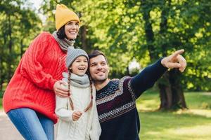 Enjoying good atmosphere. Family harmony. Happy family walk in green park, have happy expressions. Affectionate father points with fore finger into distance as tries to show beautiful sunset photo