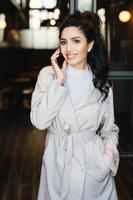 Vertical portrait of elegant brunette businesswoman in white coat communicating over cell phone while standing outdoors. Caucasian woman with beautiful appearance using modern gadget. Fashion concept photo