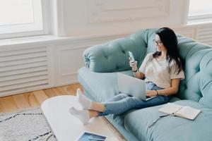 Horizontal shot of woman freelancer earns money online, checks email on smartphone, surrounded by modern technologies, organizer for making notes, uses high speed internet, sits on comfortable couch photo