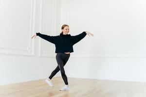 Professional female ballerina practices dance in hall, stretches hands, stands with crossed legs, has gentle smile on face, dressed in black sportswear, enjoys spare time for hobby. Healthy lifestyle photo