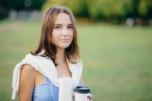 Photo of beautiful comely girlfriend with white sweater on shoulders, drinks takeaway coffee, thinks about something, stands outdoor, has walk across green field. People, lifestyle and spare time