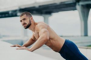 una foto al aire libre de un hombre musculoso barbudo hace ejercicios de empuje, se calienta antes de entrenar, posa contra el fondo del puente, hace ejercicio temprano en la mañana, se concentra en algún lugar. Estilo de vida activo