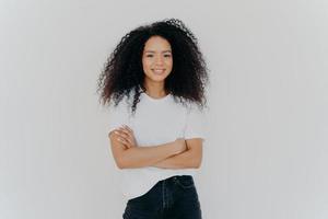 Studio shot of carefree young beautiful woman with Afro hairstyle, keeps arms folded, smiles joyfully, wears casual t shirt and jeans, isolated on white background. People, ethnicity, face expressions photo