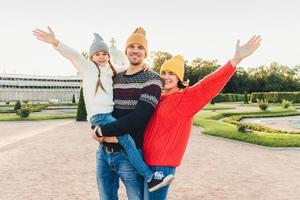 Three people have good time together mother, father and small daughter stand next to each other, walk outoors, wave with hands, have happy expressions. People, facial expressions and emotions photo