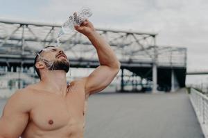 Tired sportsman cools off with fresh water poses with naked torso outdoor, tries to refresh himself, leads active lifestyle, takes break after cardio training. Excercising, refreshment concept photo