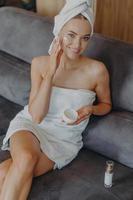 Vertical shot from above of good looking cheerful European woman applies face cream on cheek, uses cosmetic lotion, smiles gently, wrapped in white bath towel, poses on comfortable sofa at home photo