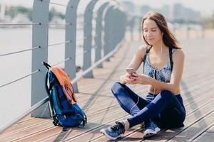 Horizontal shot of beautiful happy atthletic female in sneakers, sportswear, feels relaxed while sits outdoor, uses modern cell phone for surfing internet, dowloads music for playlist, listens song photo