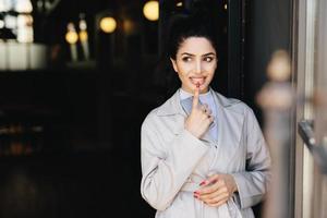 Fashionable lady with dark luminous hair tied in pony tail having nice eyes and red manicure holding her indexfinger on full lips and cell phone in hand having mysterious expression looking aside photo