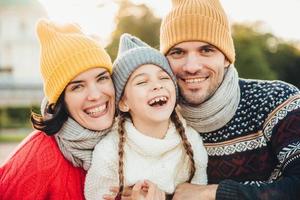 Horizontal portrait of smiling happy woman and man have good time with their daughter, wear knitted hats, warm sweaters and scarfs, going in park to have walk together. Spending time in family circle photo