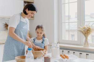 Photo of happy mum and child cook together at kitchen, wears aprons prepare something tasty, make food, whisk eggs with beater, bake house pastries at home. Milk, eggs, chocolate, flour on table