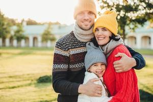 Horizontal portrait of young handsome man in yellow hat and warm knitted sweater, embrace his wife and daughter, pose at camera while stand in park. Three family members spend weekends together photo