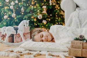 Restful small child lies on soft white pillow, sees pleasant dreams, fall asleep near decorated New Year tree, waits for presents before Christmas Eve, has tender smile on face. Children, coziness photo
