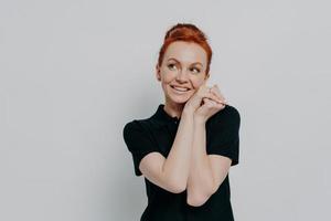 Portrait of dreamy ginger girl looking aside at copy space with smile, holding hands under chin photo