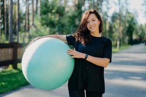 Fitness, sport and healthy lifestyle concept. Smiling curly brunette woman in black t shirt holds big fitness ball poses outside against forest background, breathes fresh air during sunny day photo