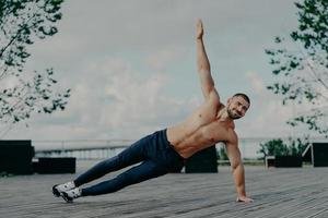 Healthy man stands in abs side plank and raises arm, does exercises outdoor, has muscular body and pleased expression. Handsome athlete trains in open air. Male couch balances on hand. Sport concept photo