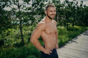 Active healthy lifestyle concept. Cheerful sporty man works out in nature, enjoys athlete exercising, keeps hands on hips, has muscular body, looks somewhere with cheerful smile does morning gymnastic photo