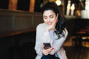 People, communication and lifestyle concept. Happy brunette woman with attractive face looking elegantly making video call, using online app on her mobile phone or listening to music relaxing indoors photo