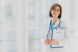 Horizontal shot of woman doctor stands with clipboard, fills up application form, holds pen, smiles positively, enjoys her work, helps people, stands in white medical gown against big window