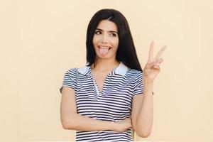 Positive funny female makes peace sign with hand, shows tongue, being in good mood, has long dark hair, has healthy skin, wears casual clothes, isolated over studio background. Body language photo