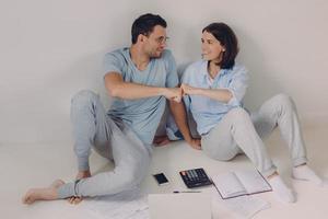 Positive successful woman and man manage finances together, give fist bump, sit on floor with papers, notebook, calculator, model over white background, agree with each other. Collaboration concept photo