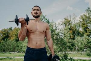 Muscular shirtless young man raises barbells outdoors, trains muscles and has strong body. Athletic sportsman with strong arms leads healthy lifestyle, enjoys training against nature background. photo