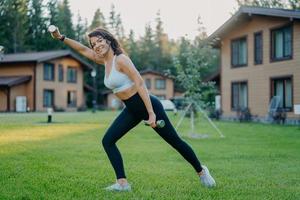 Sideways shot of active smiling determined woman raises dumbbells and has morning workout, dressed in active wear, poses on green lawn against private houses. Sportswoman trains biceps with weights photo