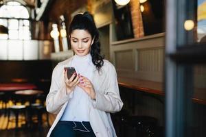 Pretty brunette woman with pony tail and appealing appearance sitting in cafe using her smartphone surfing internet using free wifi conncection and listening to her favourite music with earphones photo