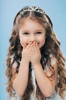 Vertical shot of positive small child covers mouth with hands, giggles positively, wears crown, expresses happiness, isolated over blue background, hears something funny. Children and emotions photo