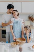 Family cook meal at home. Husband and wife embrace with love, small girl poses near, prepare delicious dinner, use milk, chocolate, eggs, make homemade pastry, pose against kitchen interior. photo