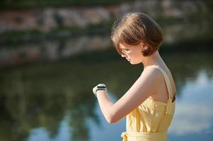 A teenage girl sets up a fitness tracker on the background of the lake. Walking tour. photo