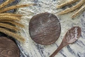 Wheat flour, wheat ear, cutting board and wooden spoon on wooden background, top view. Photo with copy space.