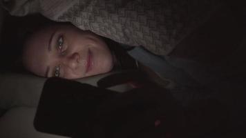 charlar debajo de la manta. mujer charlando por celular bajo la manta en el dormitorio oscuro. video