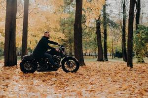 Handsome carefree bearded motorcyclist rides fast motorbike, wears shades, rides bike in park, poses against yellow trees and foliage, empty space for your information. Motor tourist outdoor photo